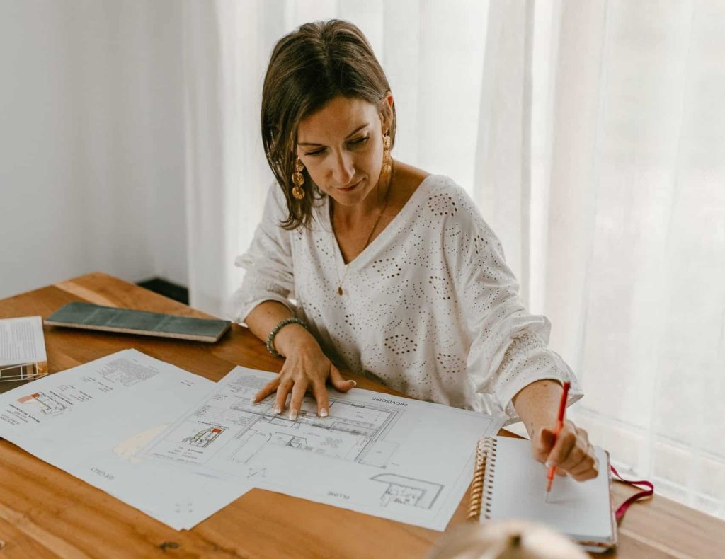 Femme qui réfléchit à l’aménagement d’un intérieur avec un plan en prenant des notes sur son cahier