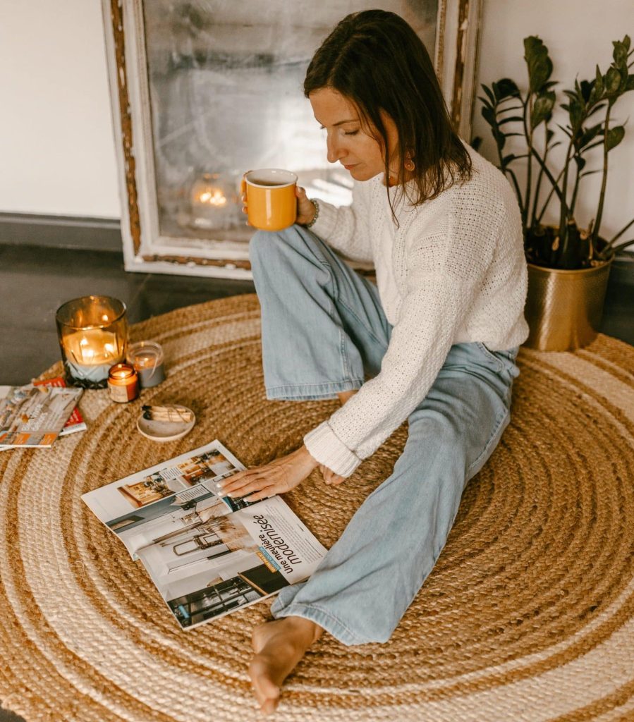 Femme assise par terre, une tasse de café à la main, entourée de bougies et consultant un magazine