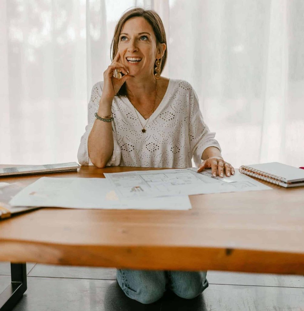 Femme réfléchissant à un projet de décoration, assise sur ses genoux avec un plan d’aménagement sur la table basse
