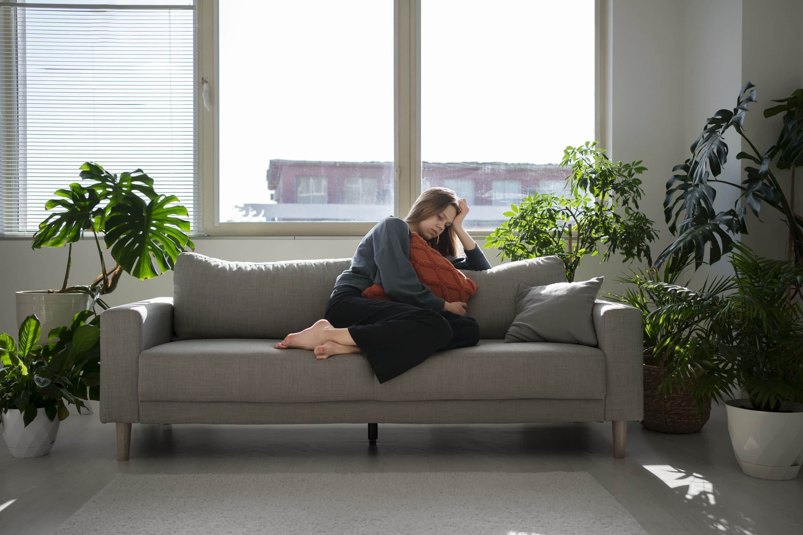 Femme triste assise sur son canapé dans une pièce entourée de plantes